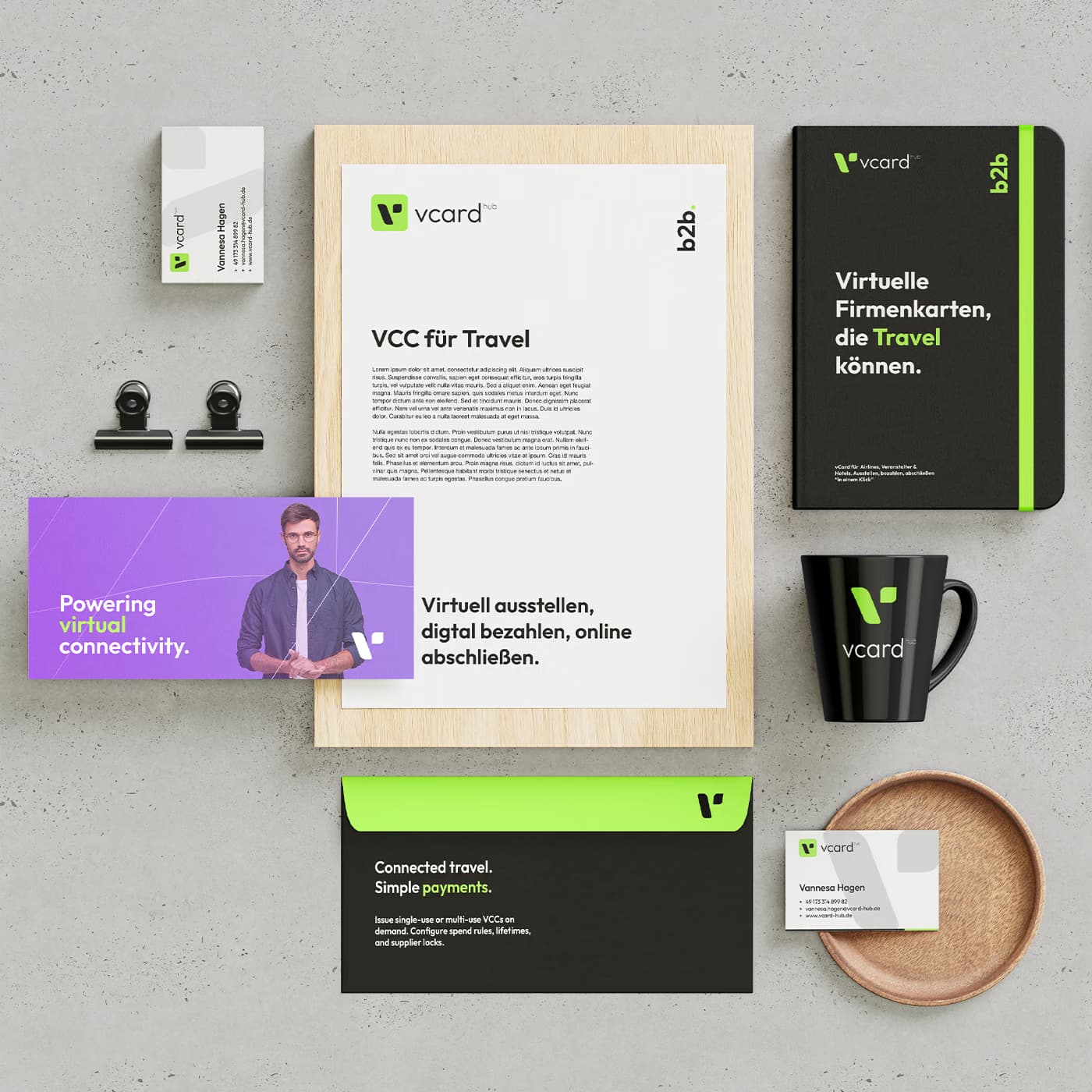 A vcard hub stationery set laid out on concrete: a letterhead headed VCC für Travel, a black notebook reading Virtuelle Firmenkarten, die Travel können, a mug, a lined envelope and business cards, in green, violet and black